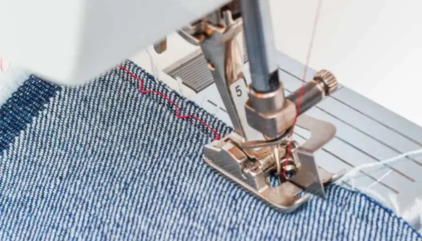 Sewing machine foot guiding denim fabric, demonstrating how to blind hem pants with a red thread stitch.
