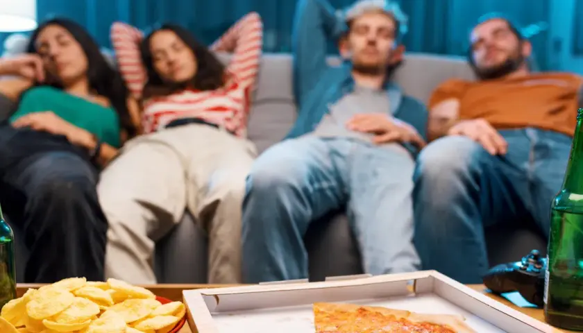 Grupa przyjaciół odpoczywa na kanapie po obfitym posiłku, czując senność. Na stole pizza, chipsy i piwo.