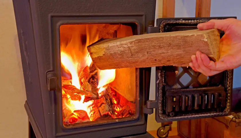Hand legt Holzscheit in brennenden Ofen. Achtung: welches Holz darf man nicht verbrennen?