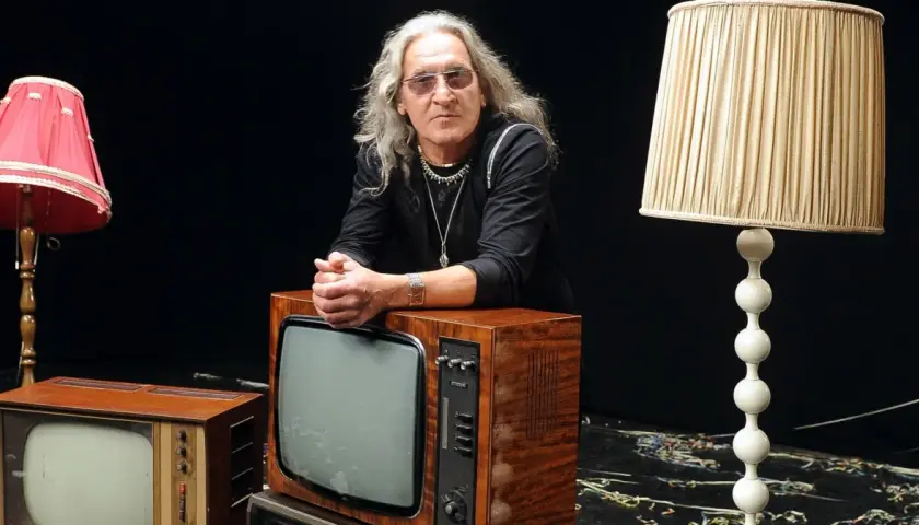 Mężczyzna z długimi siwymi włosami i okularami opiera się o stary telewizor. Wokół niego lampy i inne telewizory. Widać, że trzeba wiedzieć, kiedy ze sceny zejść.