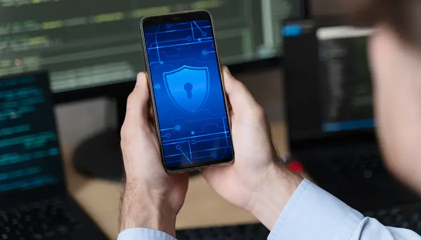 Ręce trzymają smartfon z symbolem tarczy i klucza, sugerującym odblokowanie telefonu i bezpieczeństwo danych.