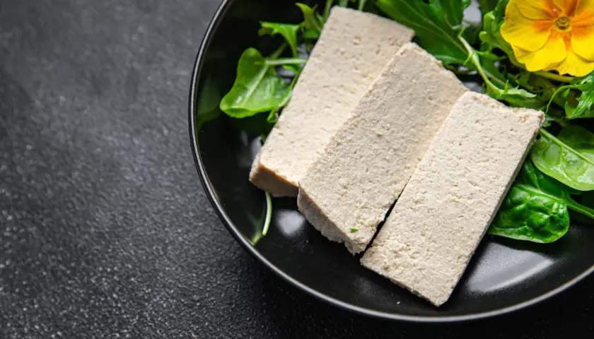 Trzy kawałki tofu na talerzu z zielonymi liśćmi i żółtym kwiatkiem. Czy tofu to nabiał? To pytanie często zadawane przez osoby na diecie roślinnej.