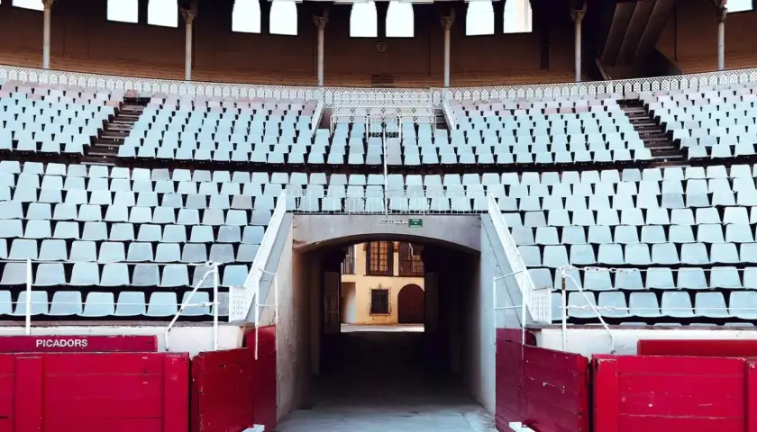 Vista de la plaza de toros vacía, con filas de asientos blancos y la contrabarrera roja.