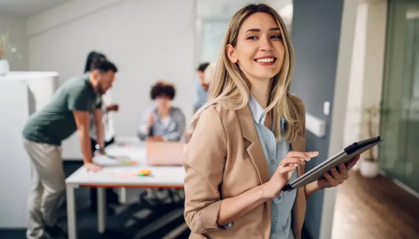 A smiling woman with a tablet, likely pursuing her masters in leadership and management, stands in an office as colleagues collaborate in the background.