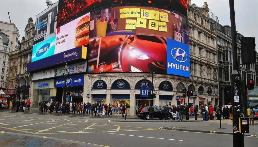 Tętniący życiem Piccadilly Circus w Londynie z neonowymi reklamami, w tym Samsung, McDonald's i Hyundai. Tłum ludzi na chodniku.