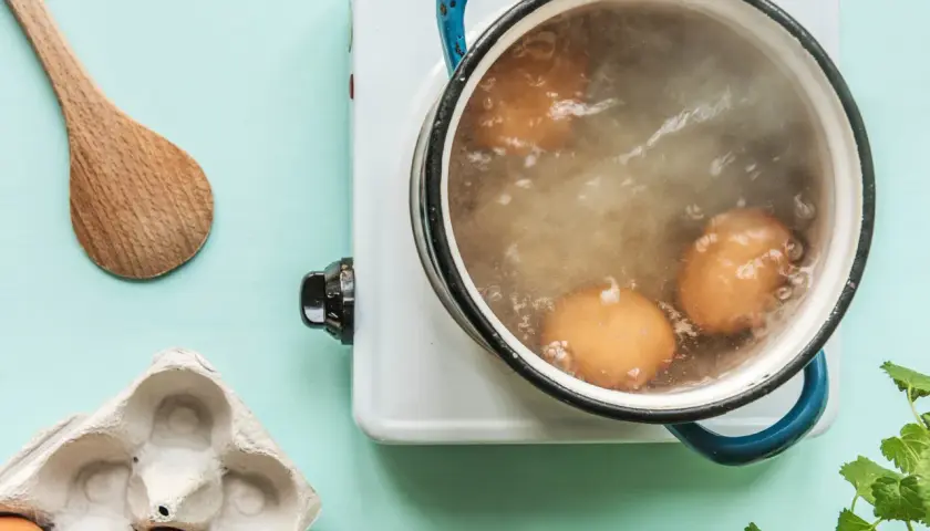 Des œufs sont en train de cuire des œufs durs dans une casserole d'eau bouillante. Une cuillère en bois et une boîte à œufs vide sont à proximité.