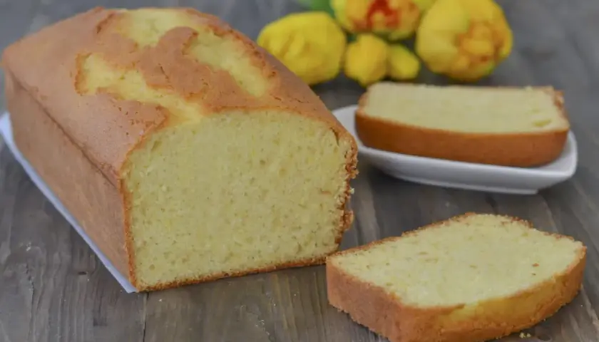Plumcake al limone soffice e profumato, con una fetta tagliata e pronta da gustare. Sullo sfondo, limoni freschi.