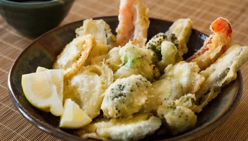 A bowl of crispy vegetable tempura, featuring broccoli, zucchini, and shrimp, served with lemon wedges and a side of dipping sauce.