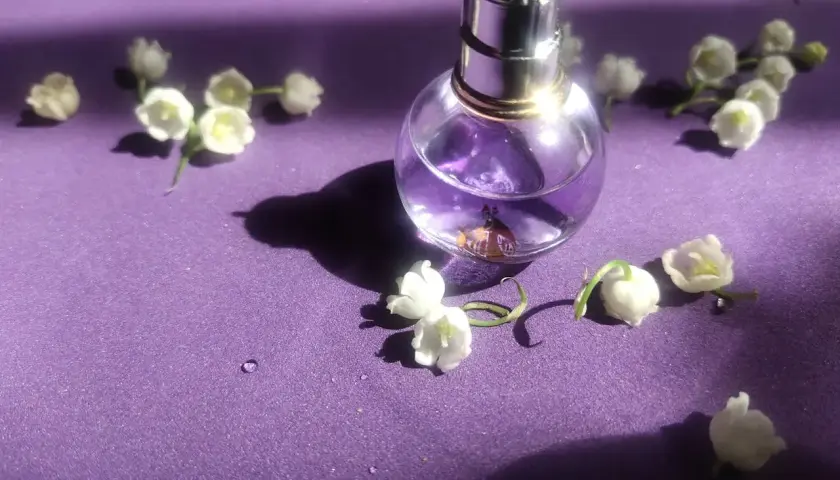 Szklana buteleczka perfum z fioletowym płynem i konwalią na fioletowym tle.