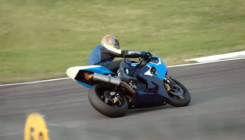 Piloto de MotoGP en plena curva, con la moto azul inclinada y llamas saliendo del escape. ¡Un auténtico holeshot!