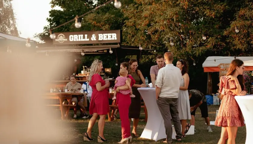 Ludzie na imprezie plenerowej przy grillu i piwie, część osób stoi przy stołach, część siedzi.