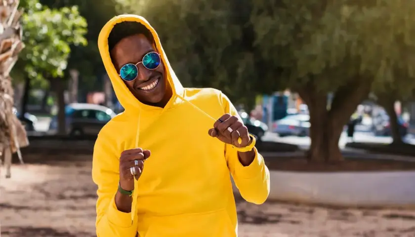 Un homme souriant porte un sweat jaune vif, ses lunettes rondes reflétant le soleil. Le grammage du sweat semble parfait pour une journée ensoleillée.
