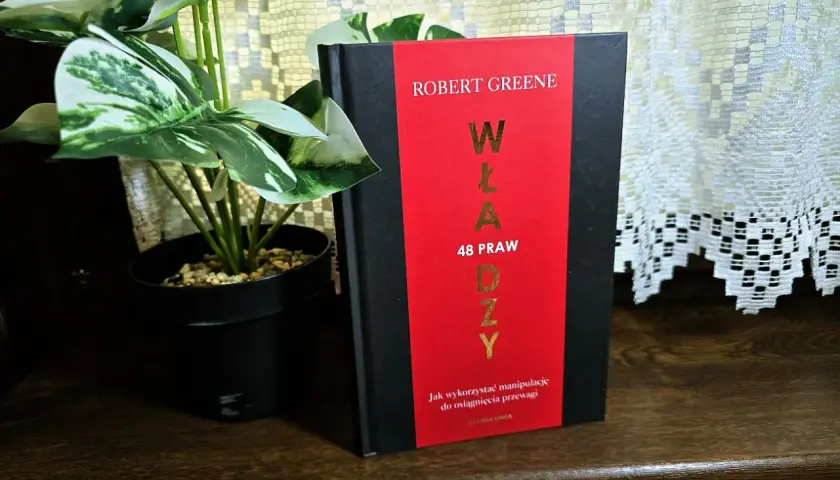 Książka "48 praw władzy" Roberta Greene'a na drewnianej półce, obok rośliny w czarnej doniczce.