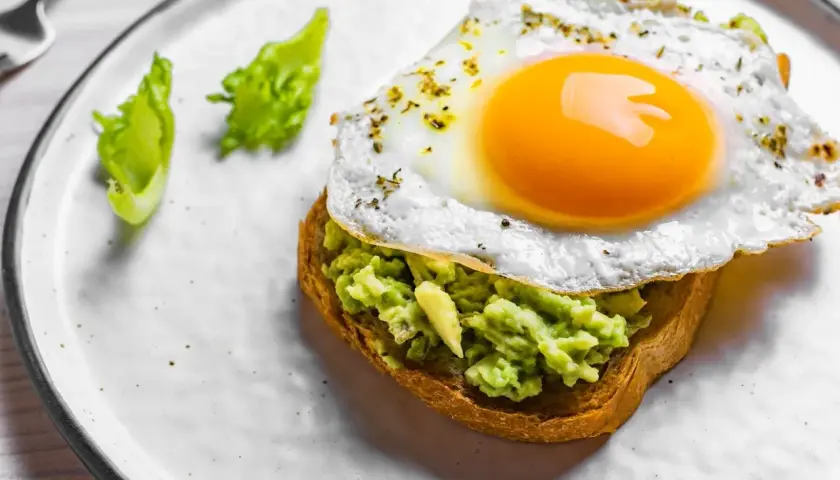 Un toast garni d'avocat écrasé et surmonté d'œufs sur le plat, assaisonné d'herbes. Deux feuilles de laitue verte décorent l'assiette.