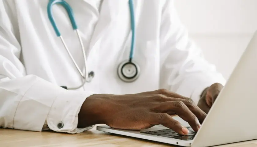 Mani di un medico che digita su un laptop, con uno stetoscopio appeso al camice bianco. Forse sta gestendo gli arretrati medici di base quando.