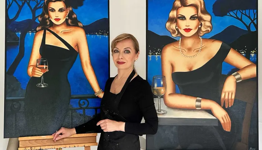 Künstlerin steht zwischen zwei Gemälden von eleganten Frauen in schwarzen Kleidern mit Gläsern Wein.