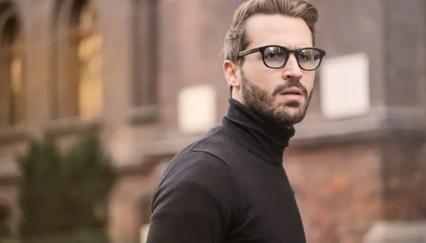 Hombre con barba y gafas, luciendo un jersey negro de cuello alto. Un estilo moderno que realza los rasgos, ideal para cortes para cara redonda hombre.