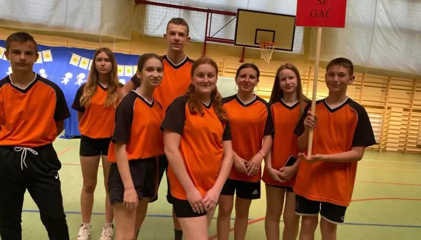 Uczniowie zespołu szkolno przedszkolnego SP GAC w pomarańczowych strojach sportowych pozują do zdjęcia w sali gimnastycznej.