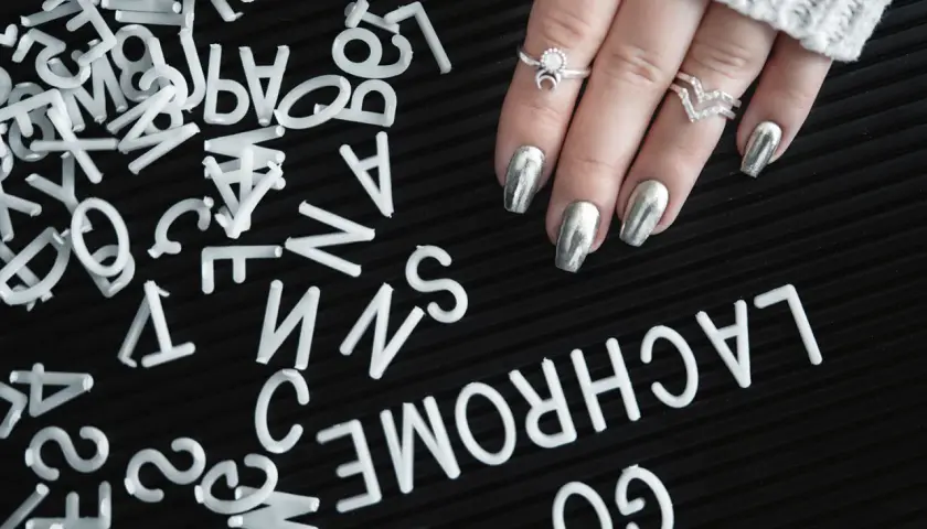 A hand with silver metallic nails and rings rests on a black surface scattered with white letters.