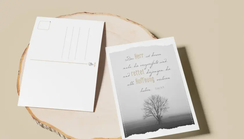 Zwei Postkarten auf einem Holzstamm. Eine zeigt einen Baum im Nebel und den Spruch: "Der Herr ist denen nahe, die verzweifelt sind und rettet diejenigen, die alle Hoffnung verloren haben.