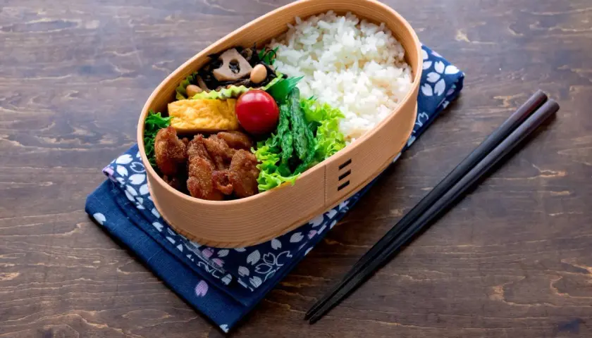 A bento box filled with rice, fried chicken, tamagoyaki, asparagus, and a cherry tomato. This is what comes in a bento box for a delicious meal.