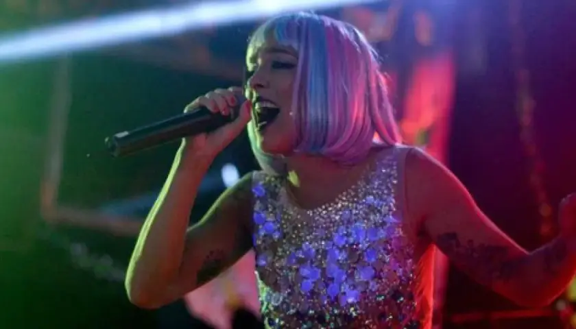 Lorena Castell, con peluca rosa y azul, canta en un escenario con un vestido brillante.