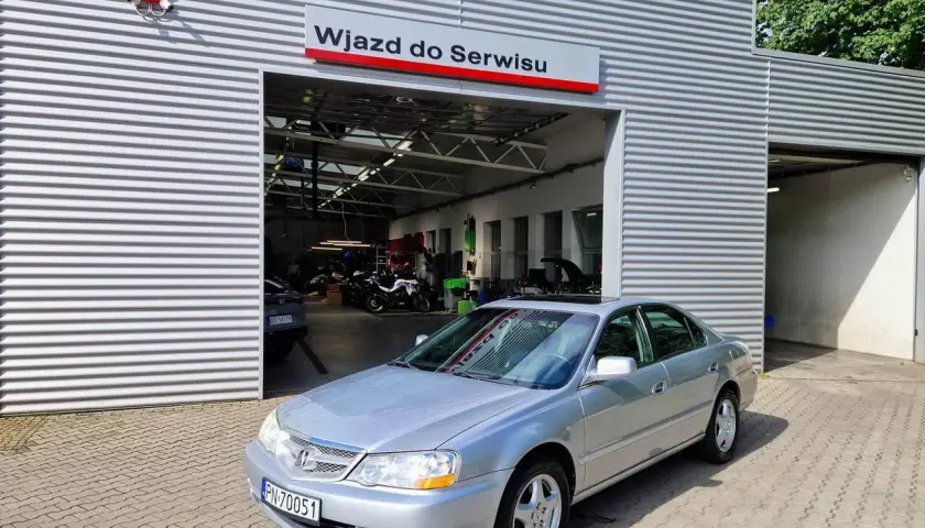 Srebrny sedan przed wjazdem do serwisu. W tle widać motocykle i sprzęt warsztatowy. Może to miejsce, gdzie naprawią mój carprog2?