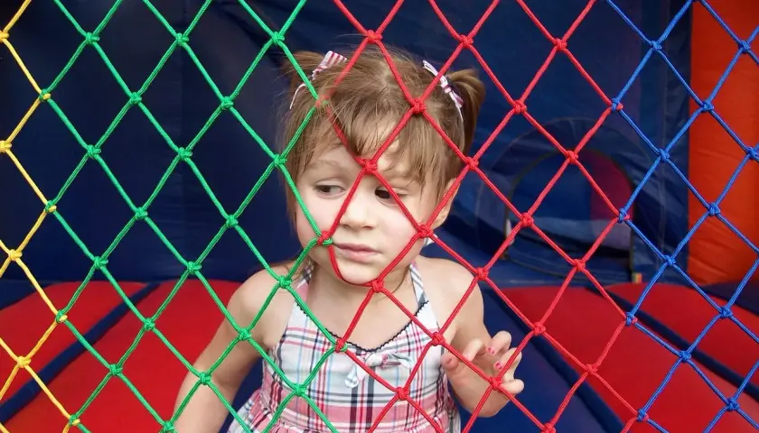Mała dziewczynka w kratkowanej sukience patrzy przez kolorową siatkę. Może słucha piosenki dla małych dzieci.