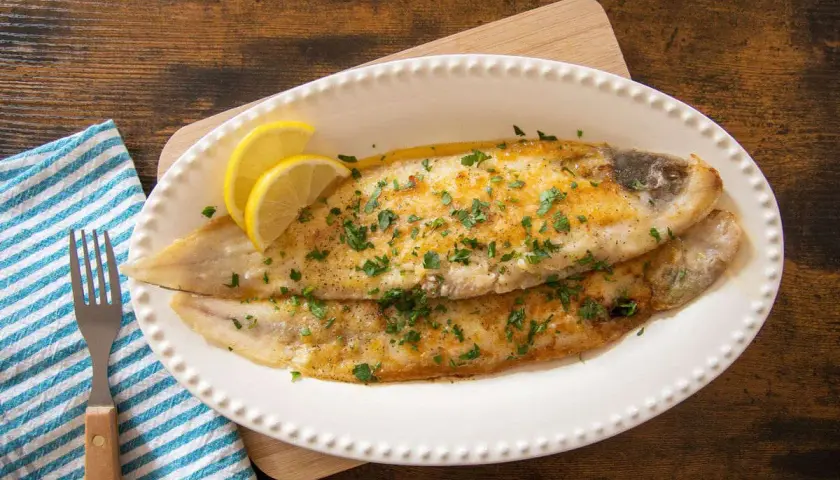 Due filetti di sogliola alla parmigiana dorati, guarniti con prezzemolo tritato e spicchi di limone, serviti su un piatto bianco.