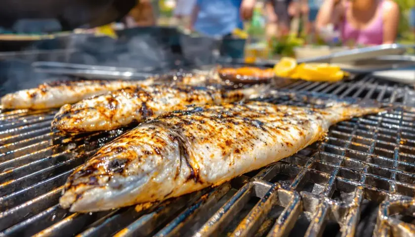 Trois poissons entiers, marqués par le barbecue, grillent sur une grille. Des éclats de citron sont visibles en arrière-plan.