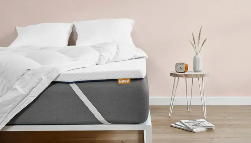 Un matelas trop mou que faire ? Ce surmatelas blanc sur un matelas gris offre une solution. Un réveil orange et des magazines sont sur une table de chevet.