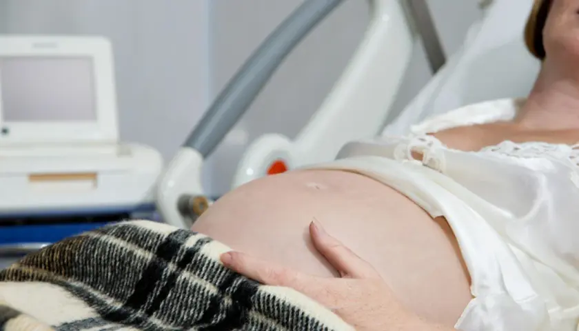 Kobieta w szpitalnym łóżku, z ręką na brzuchu, czeka na poród. Obok monitor KTG, który pomaga ocenić stan dziecka i może sugerować, jak przyspieszyć poród.