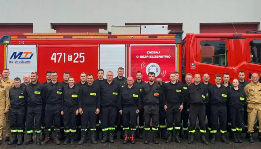 Grupa strażaków w mundurach, obok czerwonego wozu strażackiego. Dowódca sekcji OSP stoi w pierwszym rzędzie, w jasnym mundurze.