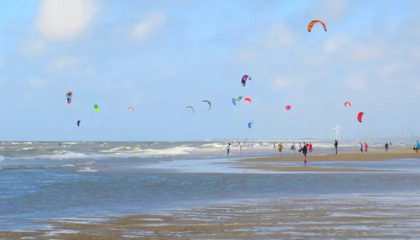 Plaża z wieloma latawcami do kitesurfingu na niebie i ludźmi spacerującymi po piasku.