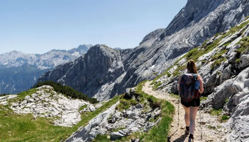 Frau mit Rucksack und Wanderstöcken genießt das Wandern in den Alpen auf einem steinigen Pfad mit Blick auf majestätische Berge.