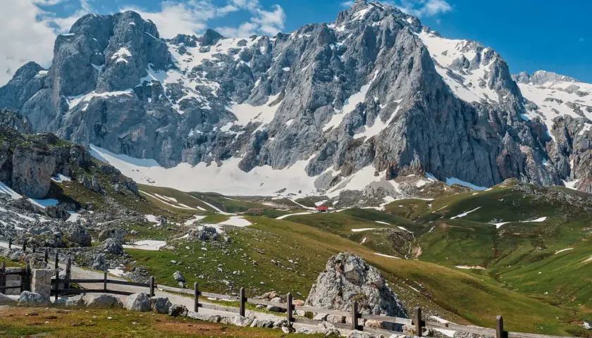 Majestuosas montañas nevadas y verdes praderas invitan al senderismo en los Picos de Europa. Un refugio rojo se asoma entre las colinas.