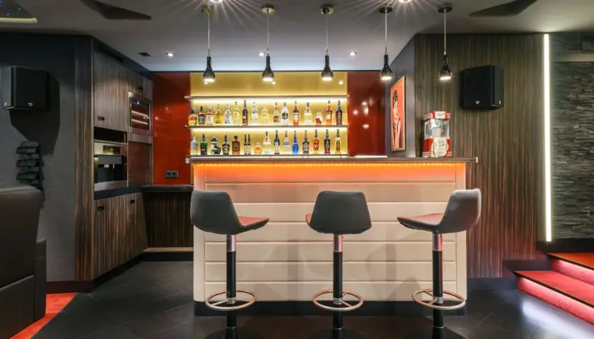 Stilvolle Bar mit beleuchteten Regalen voller Flaschen, drei Barhockern und einer Popcornmaschine. Tolle partyraum bar ideen für Ihr Zuhause.