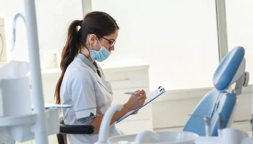 Studentka stomatologii w masce i okularach notuje coś na kartce. W tle widać fotel dentystyczny.