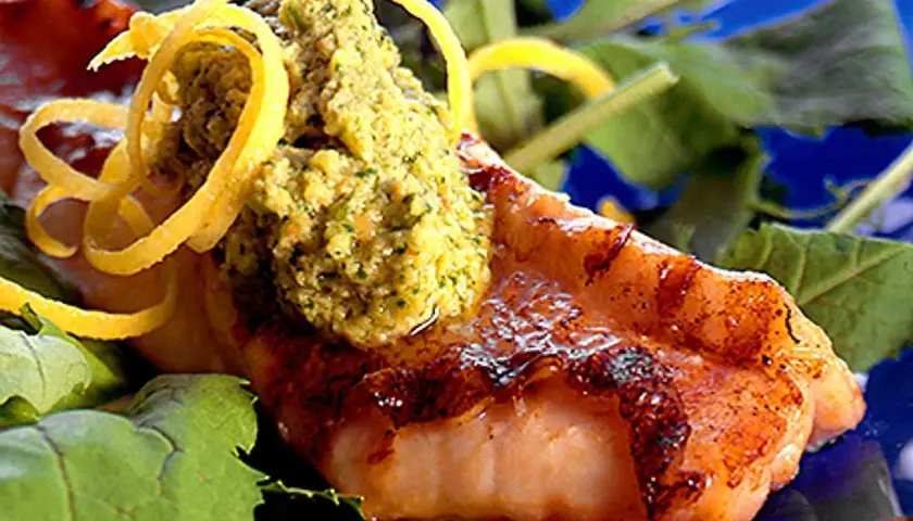 Grillad havsabborre med en grön topping och citronskal, serverad på en bädd av gröna blad.