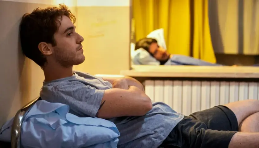 Un giovane uomo pensieroso, forse un genio incompreso o in preda alla follia, riposa su un letto in una stanza con tende gialle.