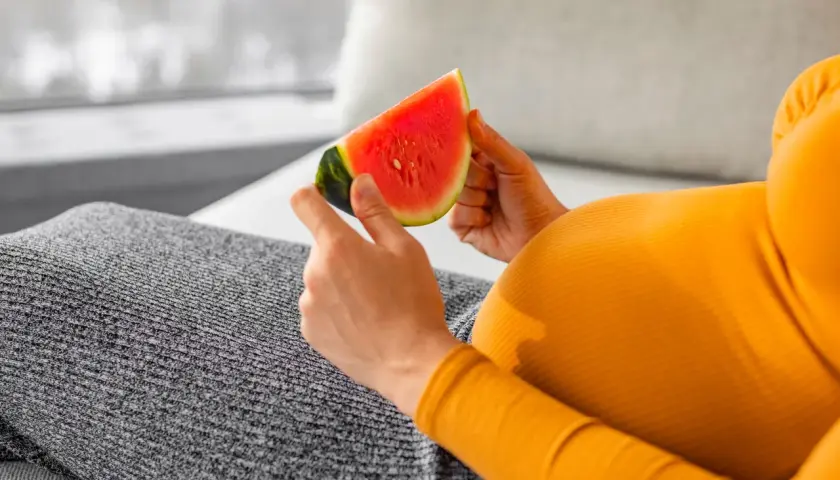 Ciężarna kobieta w pomarańczowej bluzce trzyma kawałek soczystego arbuza.