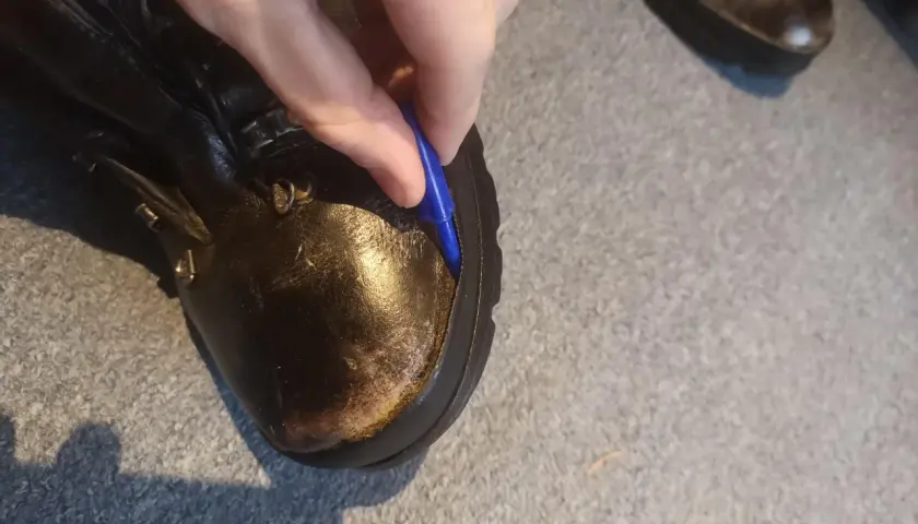 A hand uses a blue tool to pry open a seam on a leather boot, showing how to fix boots that are coming apart.
