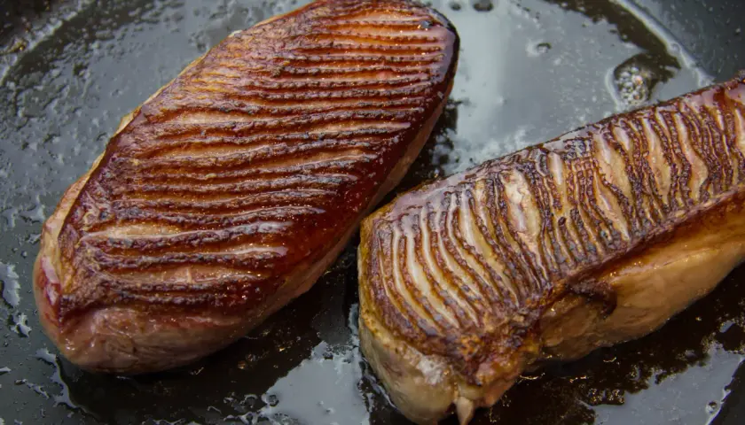 Deux magrets de canard grillés dans une poêle, prêts pour une recette savoureuse. La peau est dorée et croustillante.