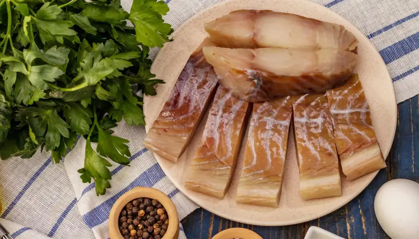 Kawałki wędzonego dorsza czarnego na talerzu, obok pęczek natki pietruszki i miseczka z pieprzem.