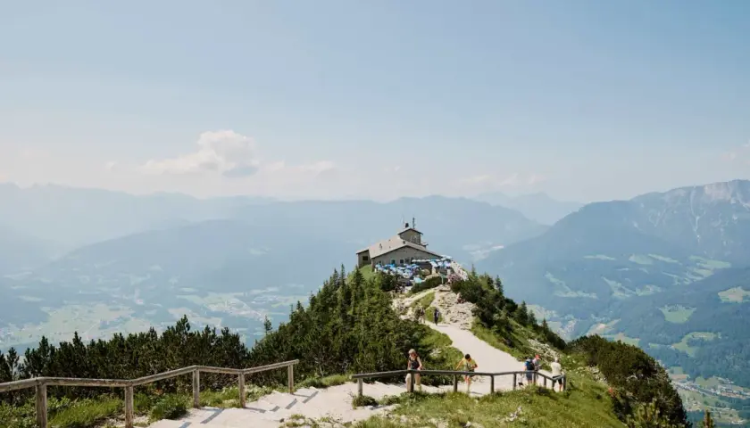 Auf einer Kehlsteinhaus Wanderung genießt man diese Aussicht: Ein Haus thront auf einem Berg, umgeben von grünen Wäldern und majestätischen Alpen.