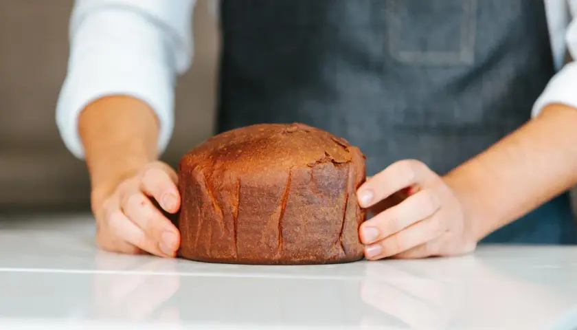 Manos sostienen un pan brioche dorado y esponjoso sobre una superficie blanca.