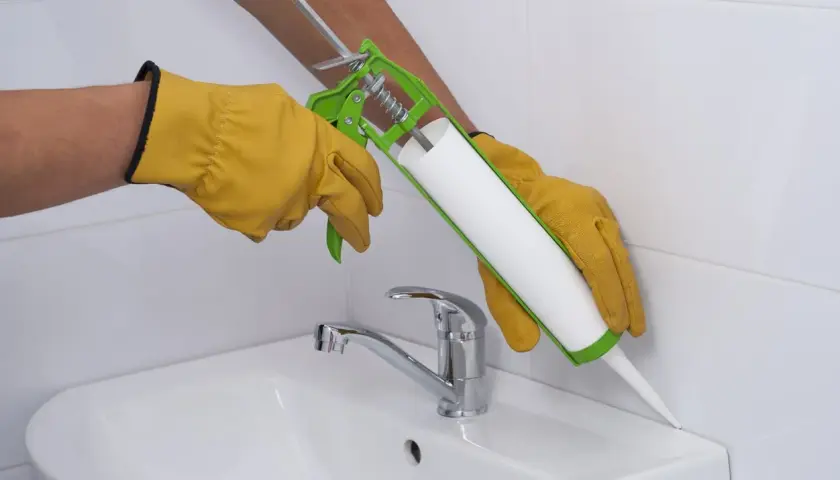 Hände mit Handschuhen dichten mit einer Kartusche den Rand eines Waschbeckens ab. Frage: wie lange muss silikon trocknen dusche?