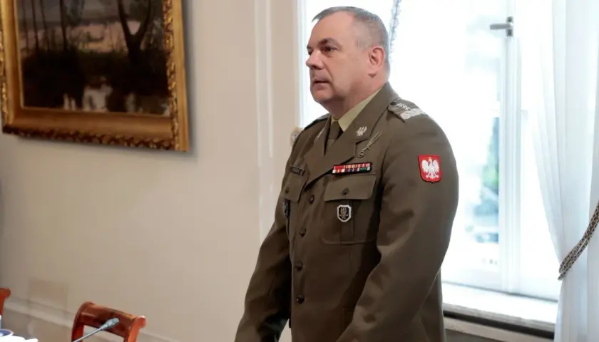 Generał w mundurze, z orłem na piersi, stoi w sali, gdzie odbywa się spotkanie korpusu generałów.