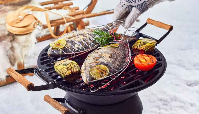 Deux poissons entiers grillent sur un barbecue, accompagnés d'artichauts et de tomates. Un gant tricoté ajuste la grille. Un vrai barbecue hivernal !