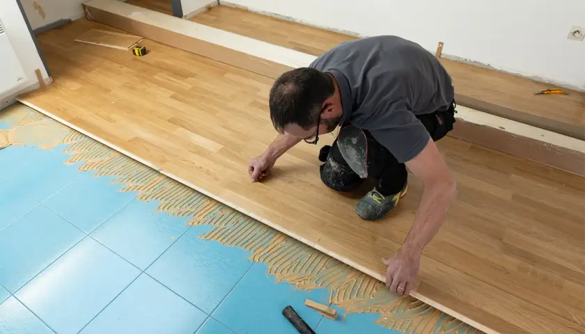 Pose d'un parquet stratifié sur un sol bleu carrelé, avec de la colle appliquée. L'homme ajuste une lame de bois.